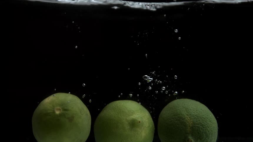 A slowmotion closeup captures three whole green limes dropping into water against a completely black background water splashes and a stream of bubbles rises to the surface as the limes settle at the bottom of the frame