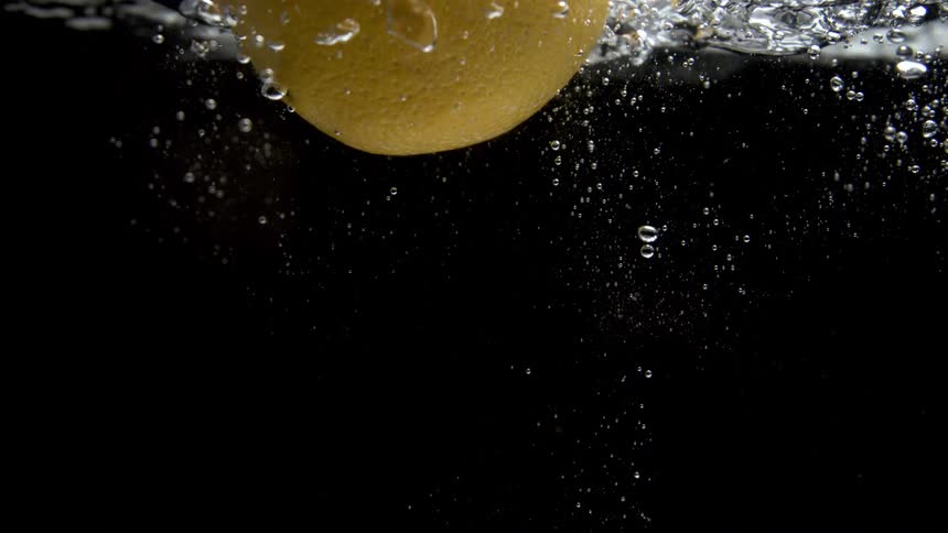A yellow citrus fruit plunges into clear water generating a cascade of splashes and a dense trail of small air bubbles that rise as the fruit descends against a uniform dark background