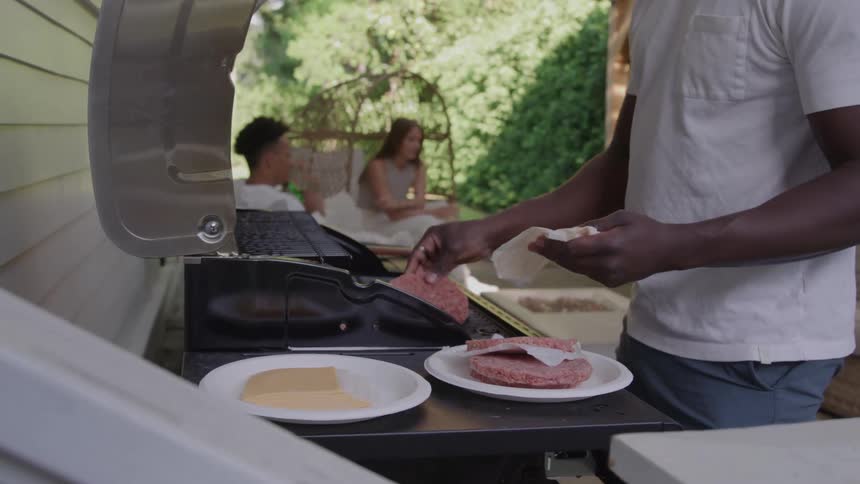 People sitting on a back patio as a man puts hamburger patties on the grill