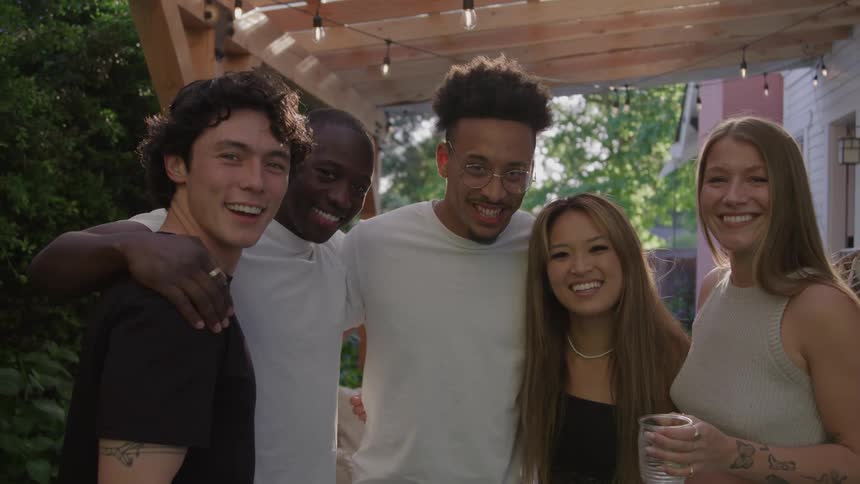A group of five friends stands close together under a wooden pergola with string lights smiling warmly as they pose for a photo outdoors in soft evening light