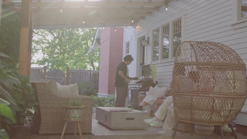 Friends relax in a patio while a barbecue is prepared under hanging lights