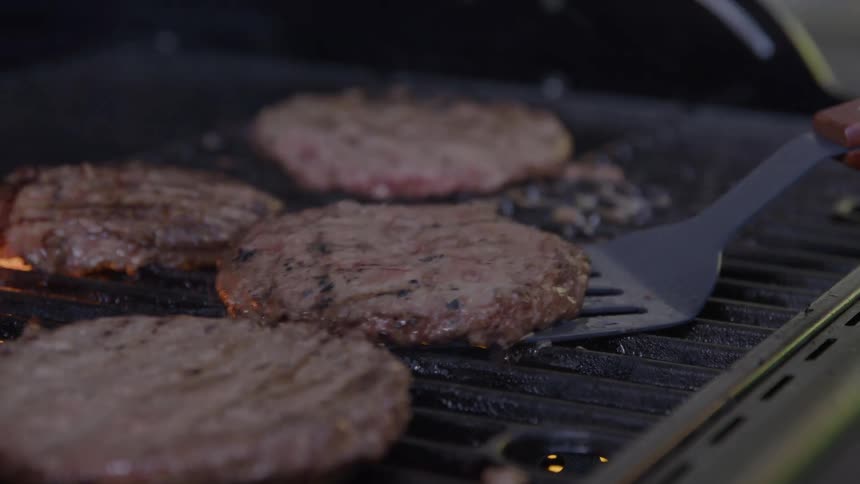 Hamburgers cook on the grill as the cook turns them over with a spatula