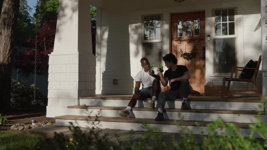 Two men sitting side by side on the porch steps of a suburban house smiling as they click their respective bottles of beer in a toast
