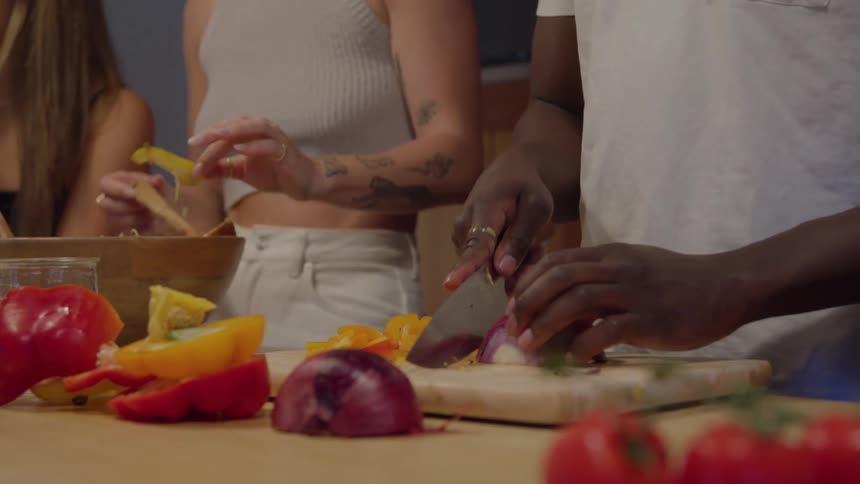 Two people are using knives to cut pieces of onions and bell peppers on a wooden board