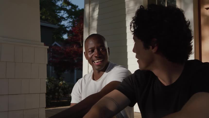 Two young men sitting on the steps of suburban houses porch talking and smiling