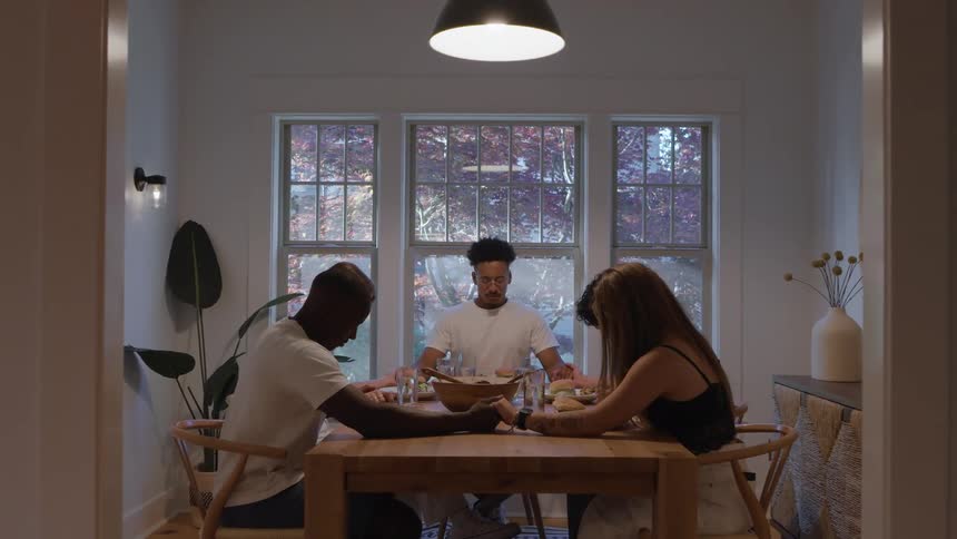 A group of friends sitting around a table in a dining room hold hands and give grace before their meal