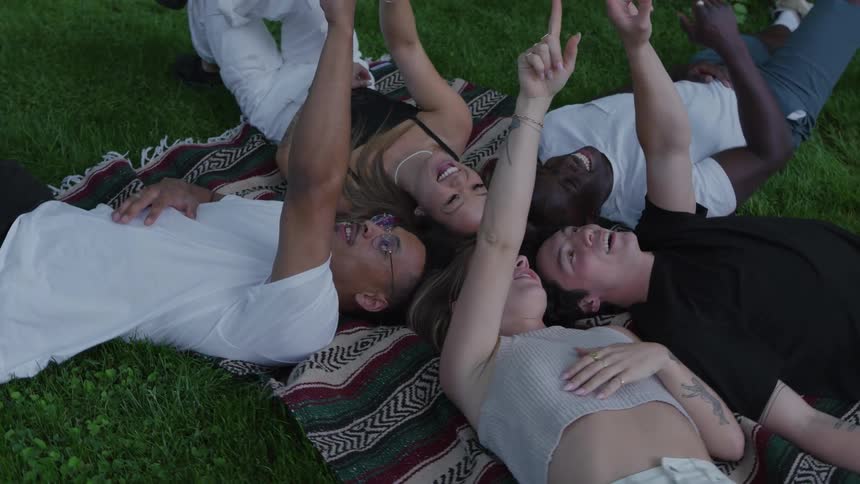 A group of five friends lying on the grass head to head in a star formation pointing at the sky while smiling and laughing