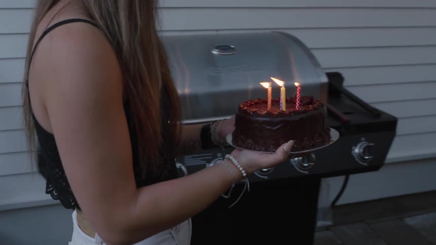 Young woman carries a birthday cake on to the patio where a man sits on the couch