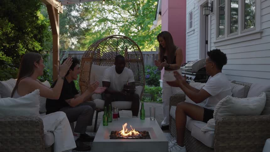A group of friends sitting or standing around a fire on a houses back porch claps and smiles as one of them holds a birthday cake and blows out the candles