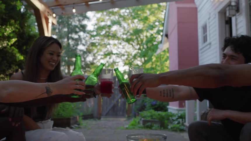 A group of friends sitting on a back porch around a fire smile as they make a toast and click their bottles and glasses together