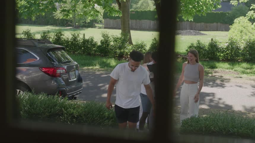 A young couple walks down the steps of a porch to greet three friends that just came out of their parked car after which they trade hugs and handshakes always smiling and walk back up to the house together