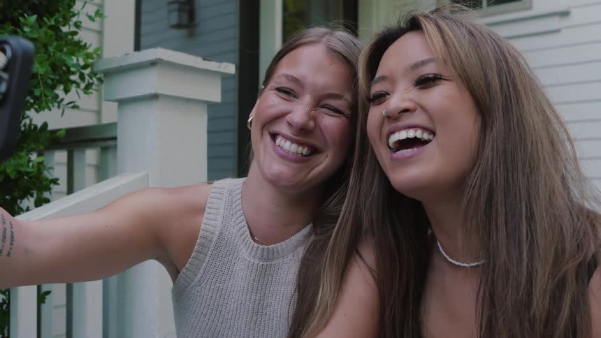 Two young women sit together on a front porch smiling as one woman holds her phone taking a selfie
