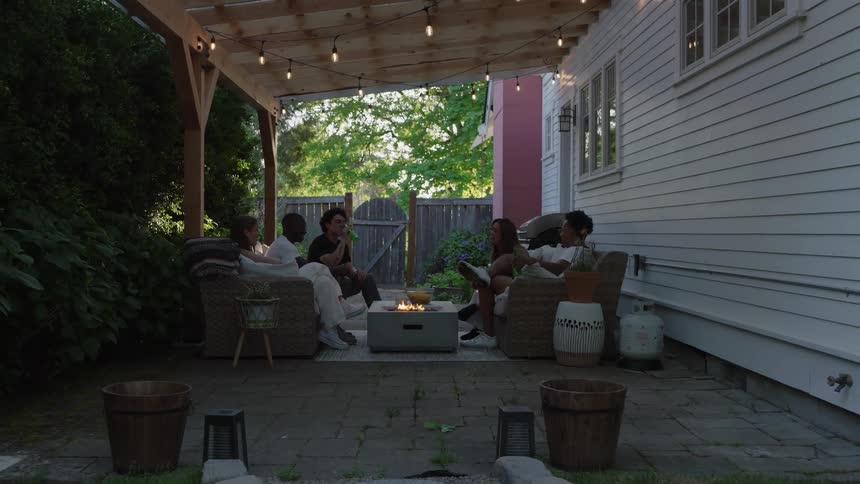 Friends relax in armchairs around a small fire pit on the patio at sunset