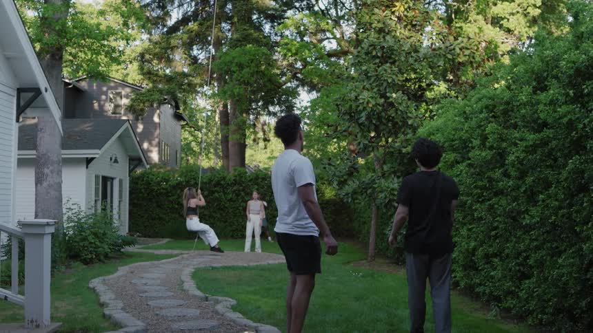 Two men are throwing a ball towards two girls who are swinging on a rope in a backyard