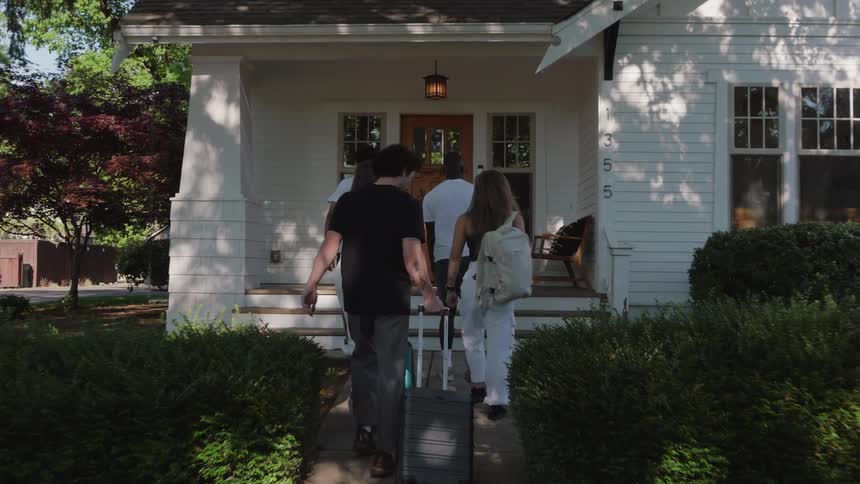 Group of people with luggage and bags walk up the sidewalk to a house