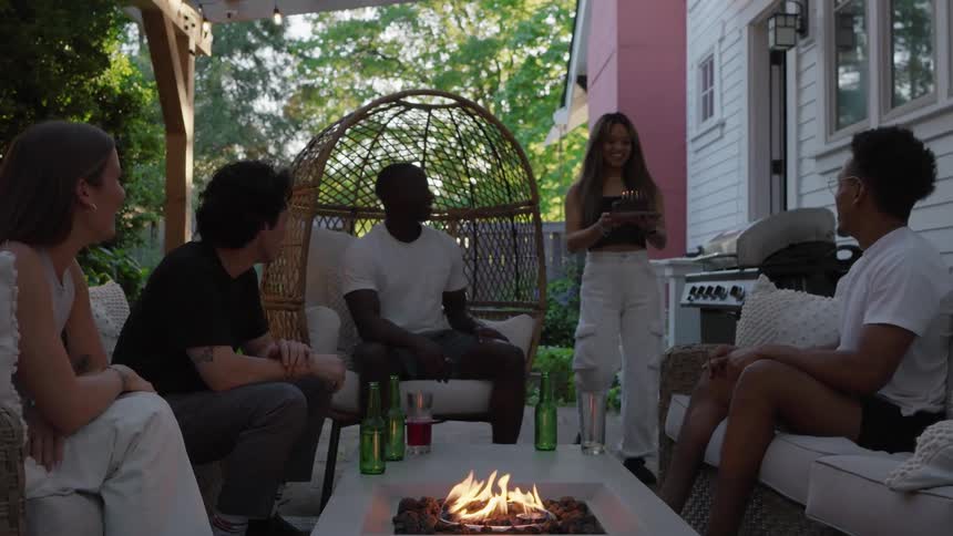 A group of friends sits around a fire on a houses back porch when a young women comes out of the house with a birthday cake and presents it to one of the sitting friends to his surprise
