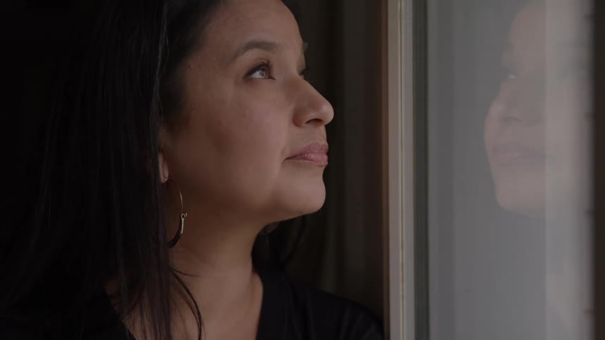 A woman with a sad expression looks out of a closed window her reflection right next to her face then she looks up