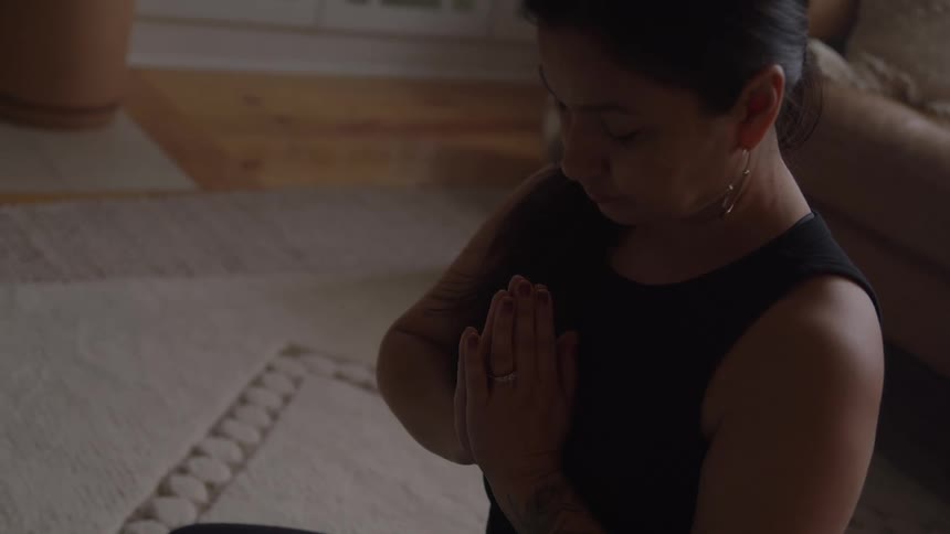 A woman sitting in a meditative pose with her eyes closed holding her hands together in the prayer position aagainst her chest within a softly lit indoor space