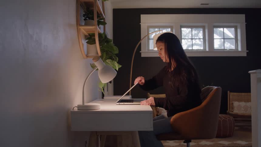 Woman sitting at a desk opens her laptop in a welllit workspace