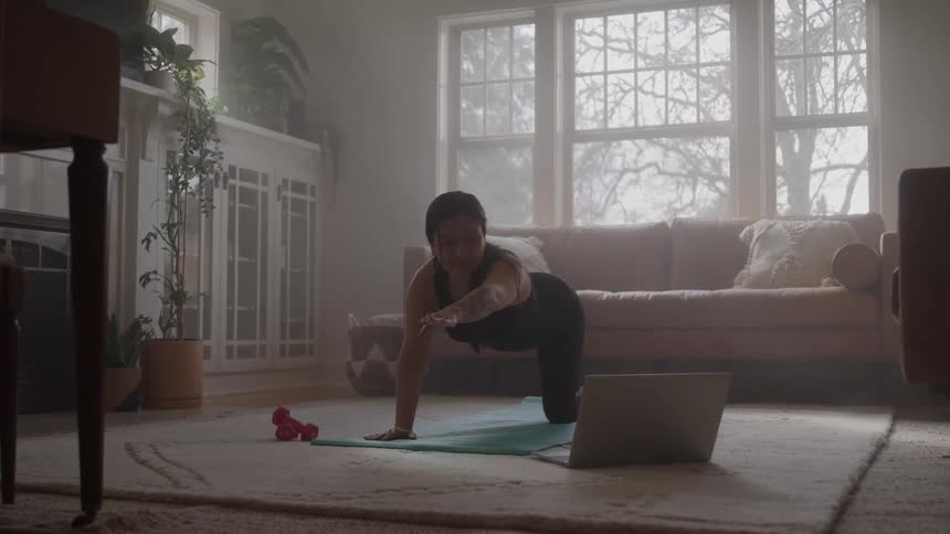 A woman on her knees on the floor of a living room doing a yoga pose while looking at the screen of a laptop on the floor next to her
