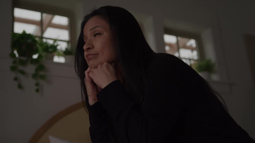 Woman with long dark hair sits with her hands over her eyes and looks up with her chin resting on her hands