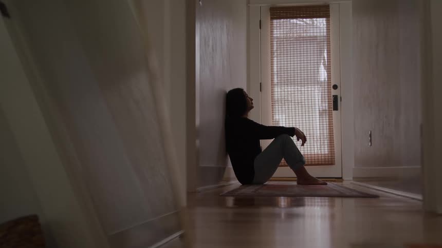 Woman sits on the floor against the wall gazing thoughtfully toward the door