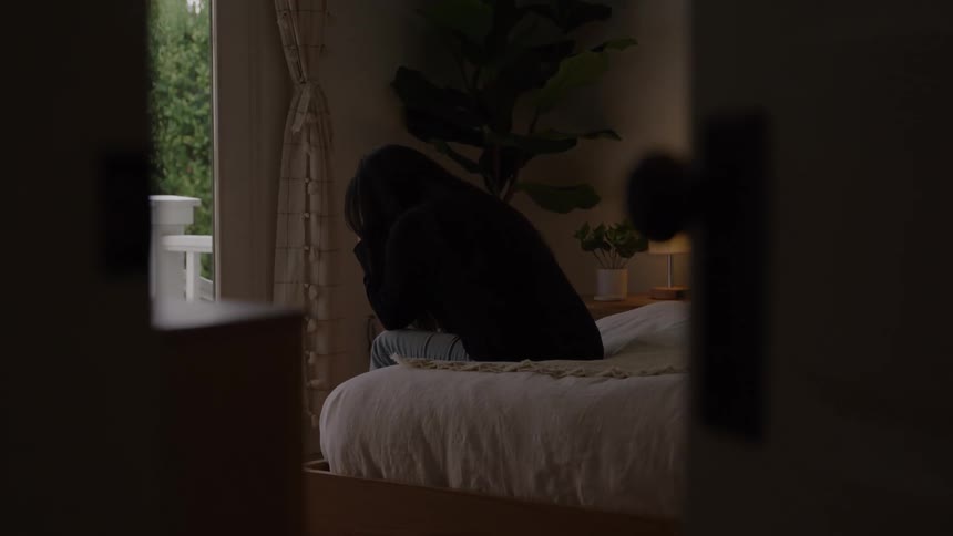 A woman sitting on the edge of a bed with her fingers interlocking bends forward and brings her hands to her face to cry