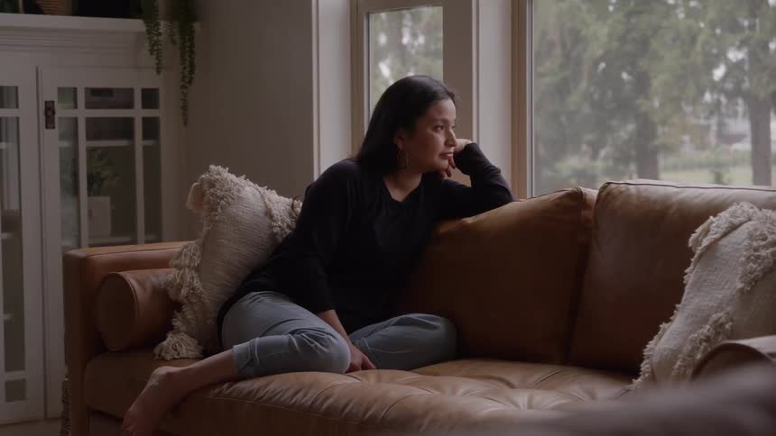 Woman sitting on her couch stares out the window while thinking