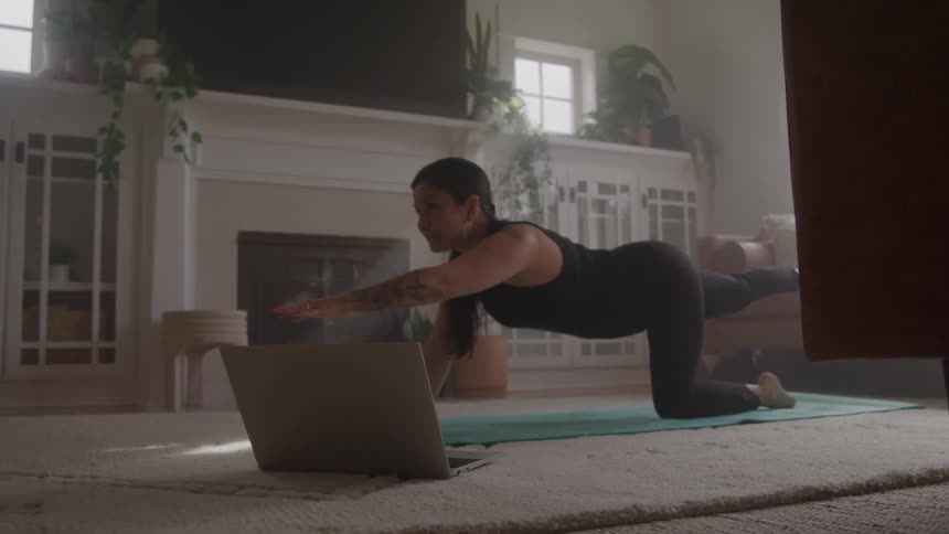 A woman performs stretching exercises in her living room next to a laptop computer
