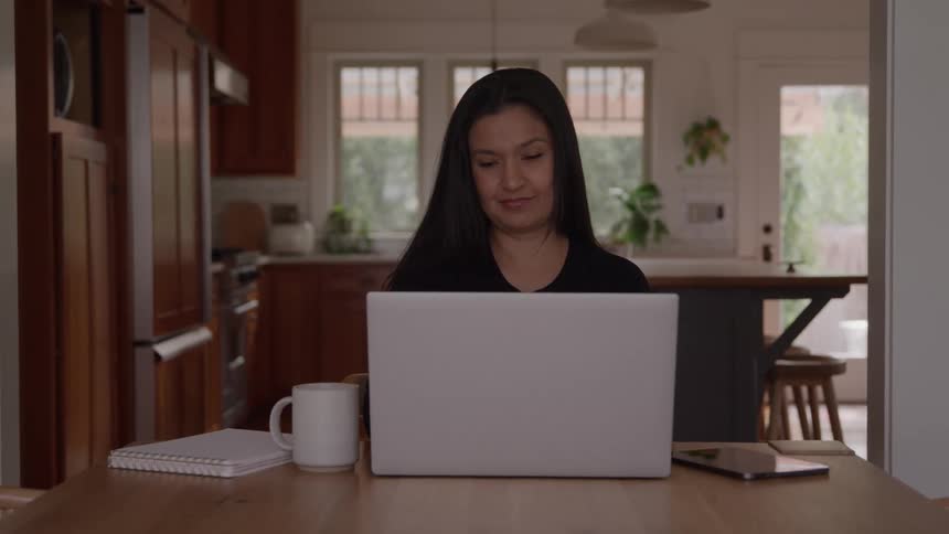 Woman works from home writing in a notebook beside her laptop