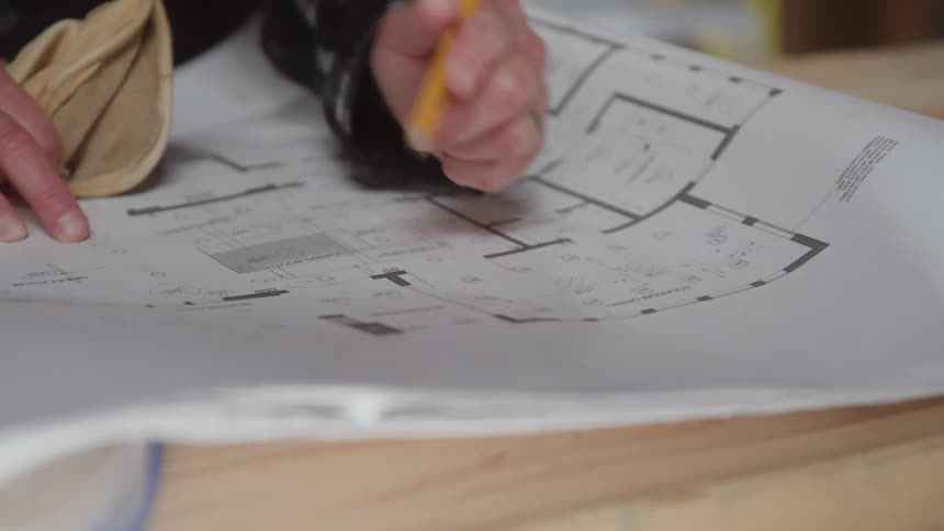 Two hands on a blueprint on white paper one of them holding a pencil and marking the blueprint