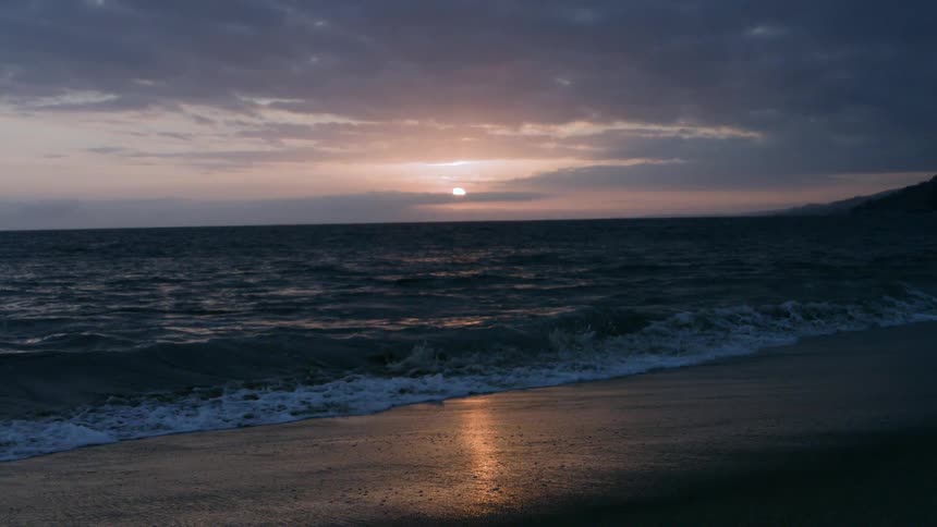 Waves roll into shore and over the beach as the sun shines through clouds on the horizon