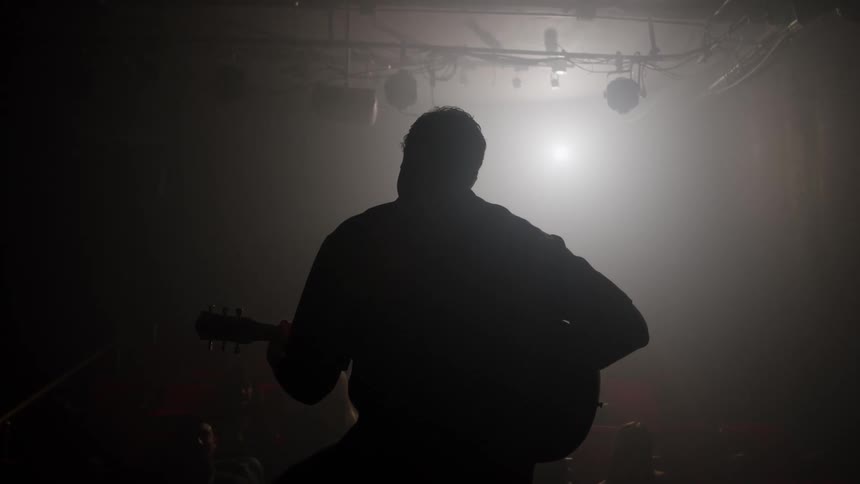 Musician plays guitar on stage under a spotlight surrounded by smoke
