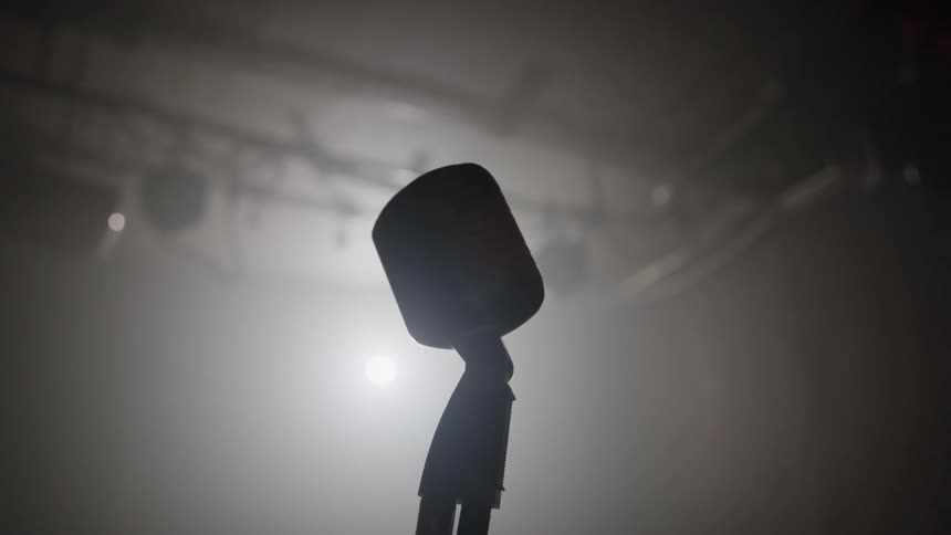 The silhouette of a vintage microphone on a stage framed by a spotlight from the ceiling