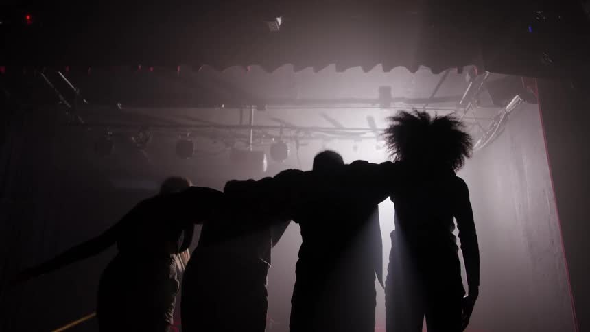 Friends with arms around each other walk toward the light on a smokelit stage