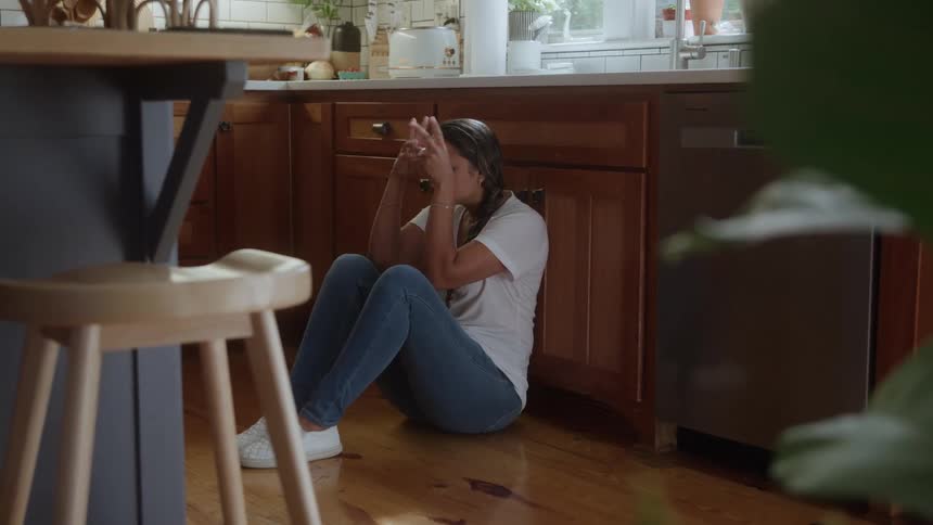 A woman in a white shirt is sitting on a wooden floor in the kitchen with her hands on her head and looking sad