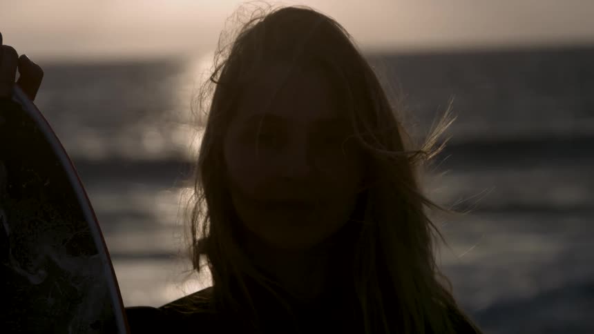 Female surfer stands on the beach backlit by the sun and holding an uprightsurfboard