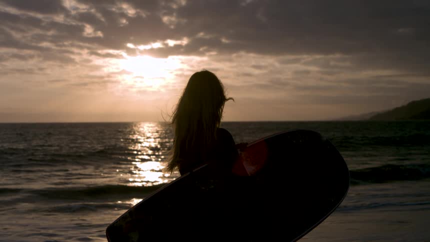 The woman holding a boogie board on the beach watches the sun rise
