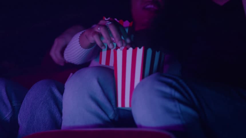 Two people eat popcorn while watching a movie in a dimly lit theater