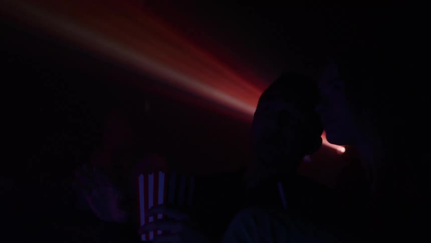 A man and woman sit in a dark movie theater watching a film illuminated by the projector beam as the man eats popcorn