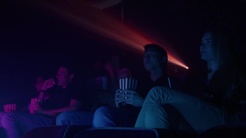 A diverse group of people seated closely in a darkened movie theater auditorium illuminated primarily by the fluctuating colorful light from the screen and the prominent dustfilled beam of the projector passing overhead and eating popcorn