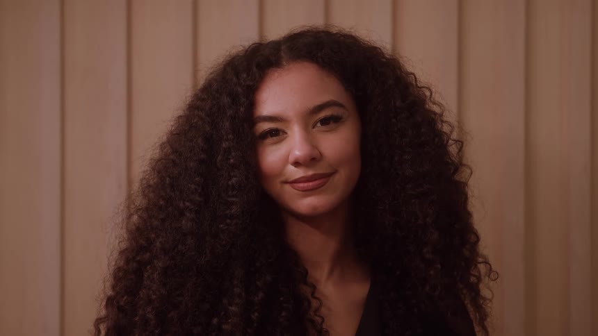 The face of a young woman who is smiling while looking ahead and she has long curly hair