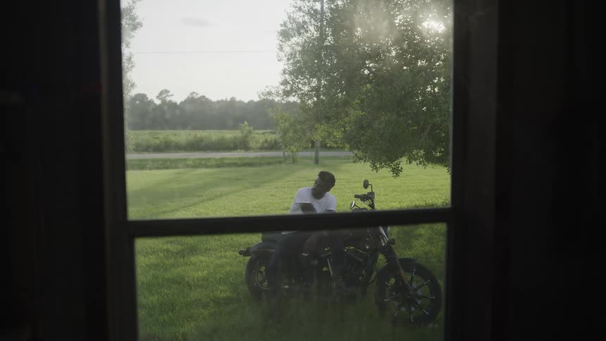 Viewed through a window a man sits on a black motorcycle parked in a grassy field on a sunny day