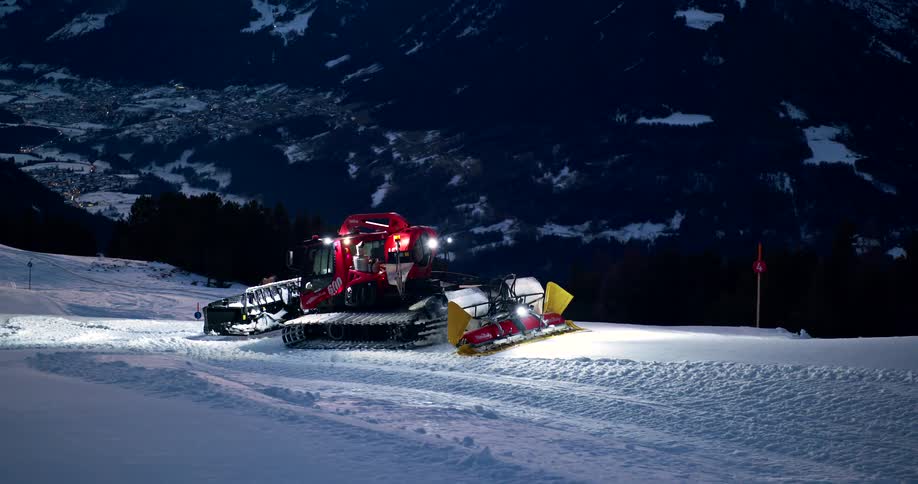 snow groomer with its lights on works on a mountain slope overlooking a lit valley at dawn