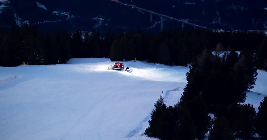 A snowmobile rides down a snowy mountain lined with trees at sunset