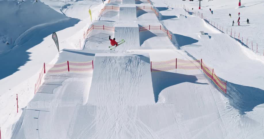 skier twists and turns as he flies over a jump in a ski park, while other skiers glide down the snow-covered slope