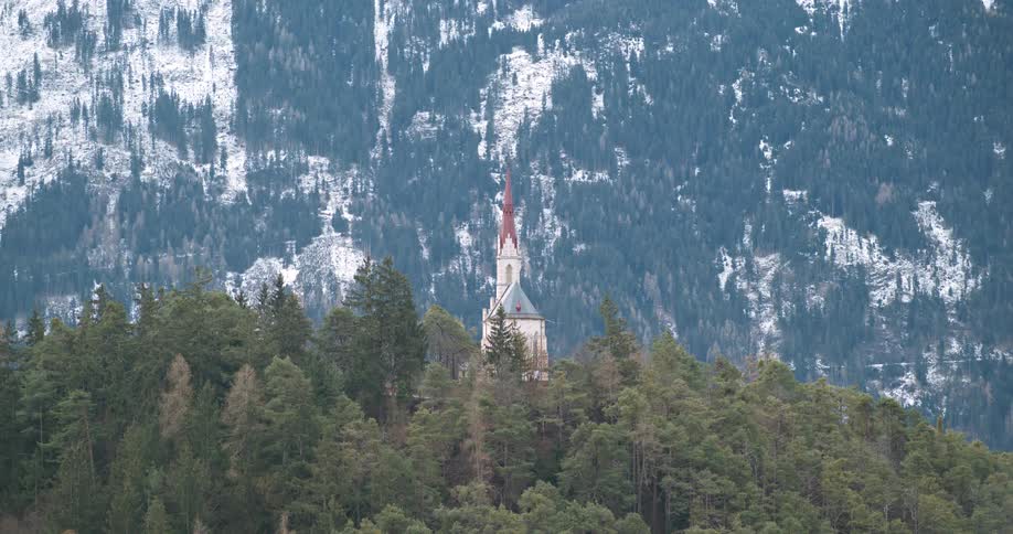 The clip offers a picturesque view of a european alpine landscape a small white stone church structure is visible just above the evergreen treetop canopy in the middle ground drawing the eye to its tall narrow spire the background is dominated by a m