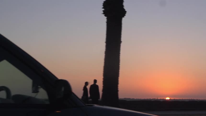 Two people walking along a sidewalk during sunset as a car drives by in the foreground