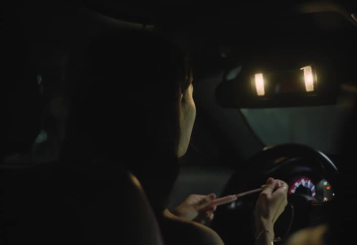 A woman sits in the drivers seat of a car at night using the lighted visor mirror to apply makeup before smiling