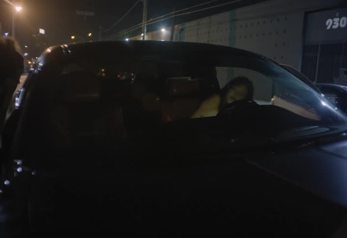 Two women are seen quickly entering and sitting in a dark vehicle parked on a dimly lit street at night with the frame dominated by the cars dark windows and the background showing a urban building under street lamps